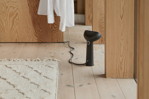 Black handheld steamer on a wooden floor with a mirror reflecting the room.